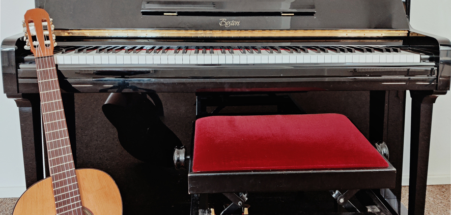 Eine Konzertgitarre lehnt an einem offenen Klavier, im Vordergrund ein roter Klavierhocker.